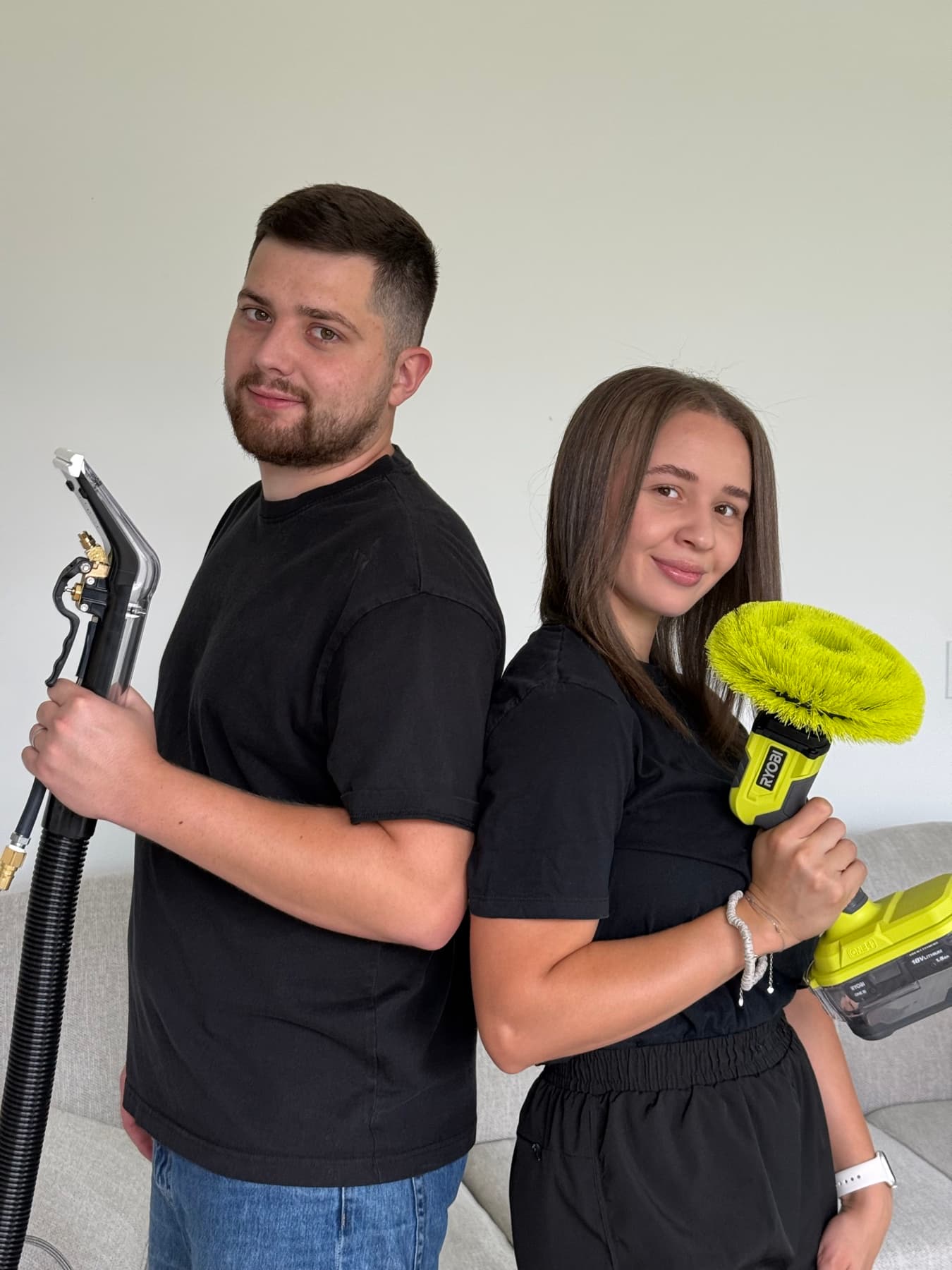 Taras and Rachel, the owners of Mr & Mrs Steam, standing back to back holding a steam extraction wand and a powered upholstery brush